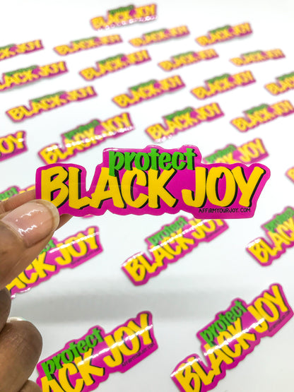 Holding a sticker that reads "Protect Black Joy" in green and yellow letters with a pink background